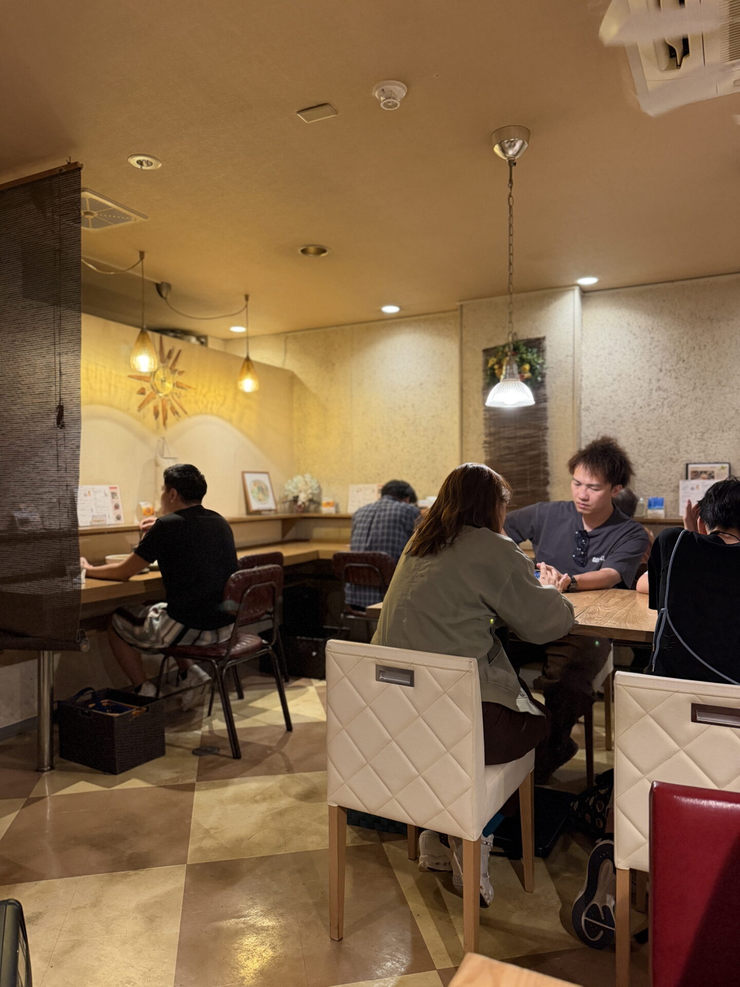 Warmly lit restaurant interior with diners sitting at wooden tables and counter seats beneath hanging pendant lights. Cream coloured upholstered chairs, patterned flooring and softly textured walls create a relaxed dining atmosphere.