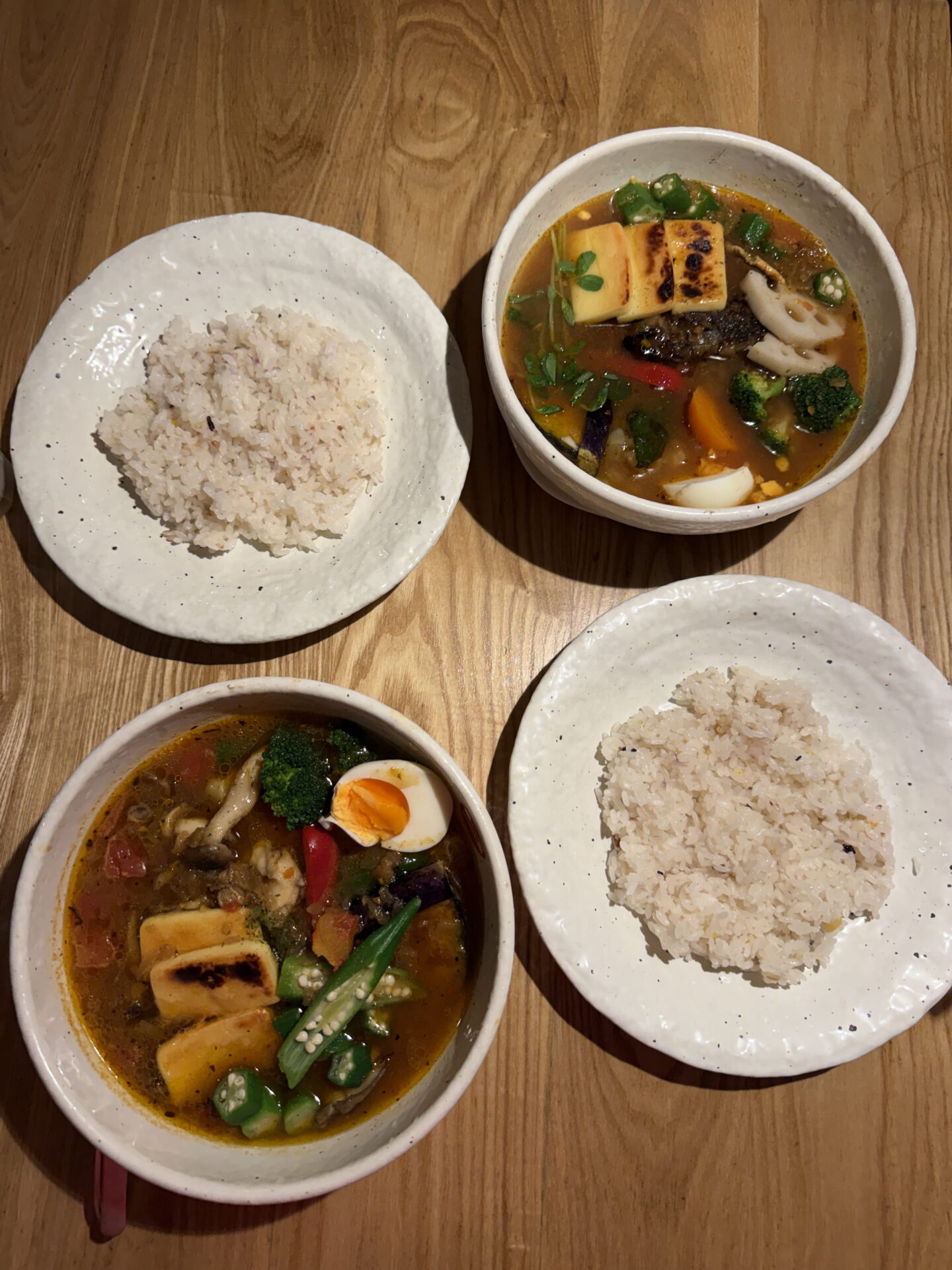 Overhead view of two bowls of Japanese soup curry served with separate plates of rice on a wooden table. The colourful soups contain grilled cheese, okra, broccoli, mushrooms, carrots and boiled eggs in a reddish brown broth.