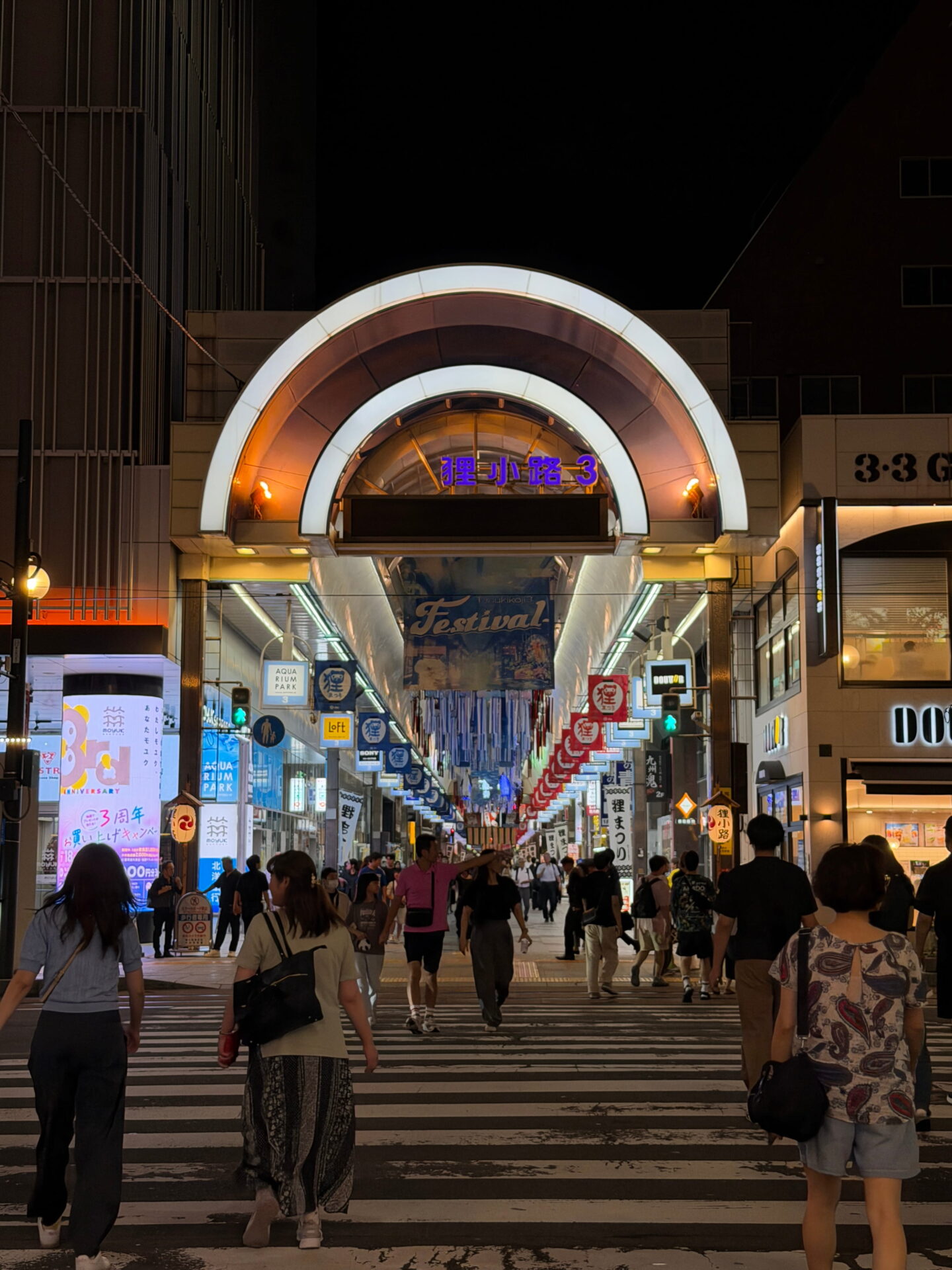 The brightly illuminated entrance to Tanukikoji Shopping Arcade 3 in Sapporo at night with its distinctive curved archway and purple Japanese lettering. Shoppers cross the road towards the covered pedestrian street lined with shops and hanging festival decorations.
