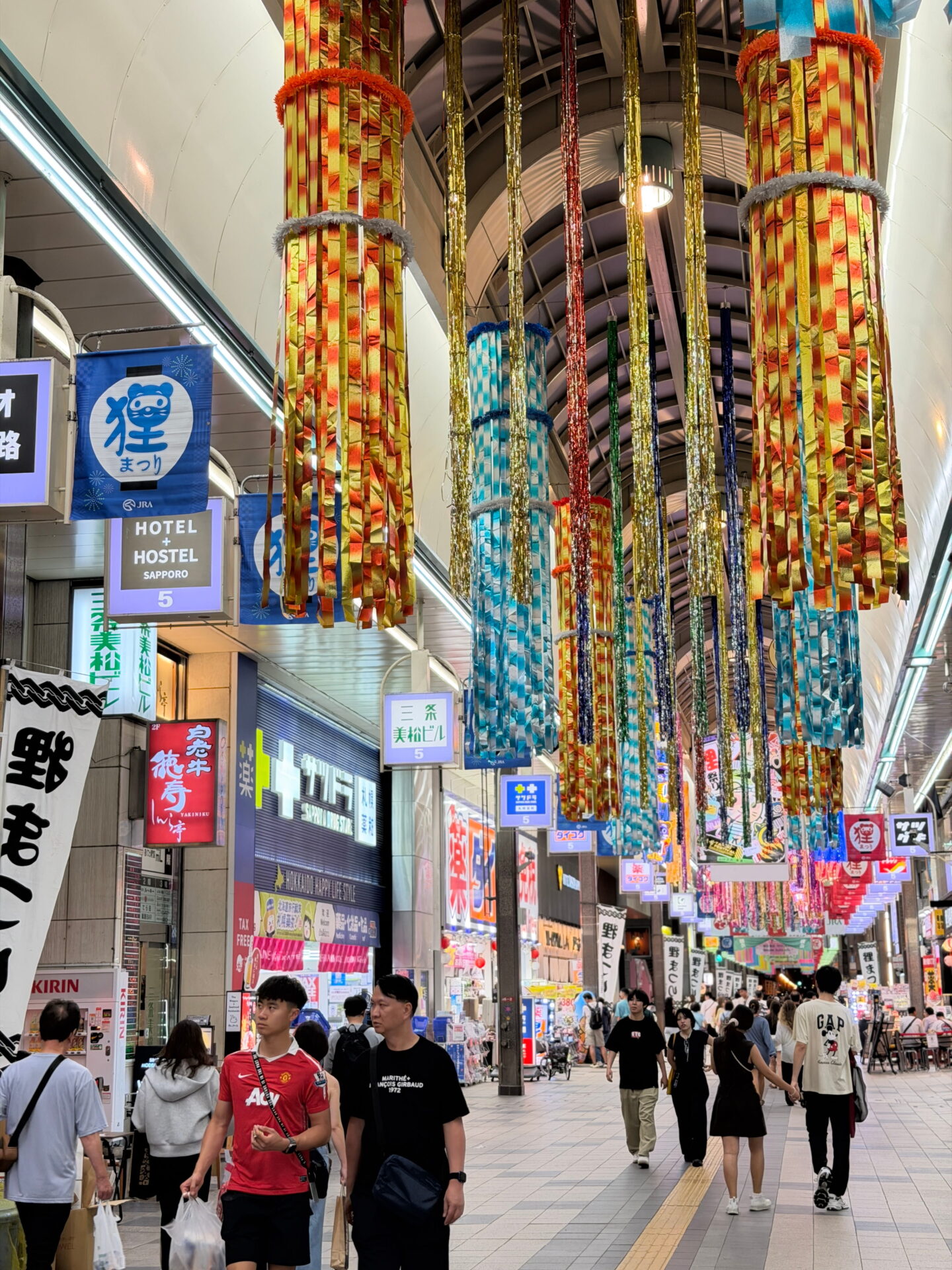 Colourful orange, yellow, blue and gold streamers hang from the high arched ceiling of Tanukikoji Shopping Arcade in Sapporo. Shoppers walk beneath the elaborate festival decorations past illuminated shopfronts and signs.
