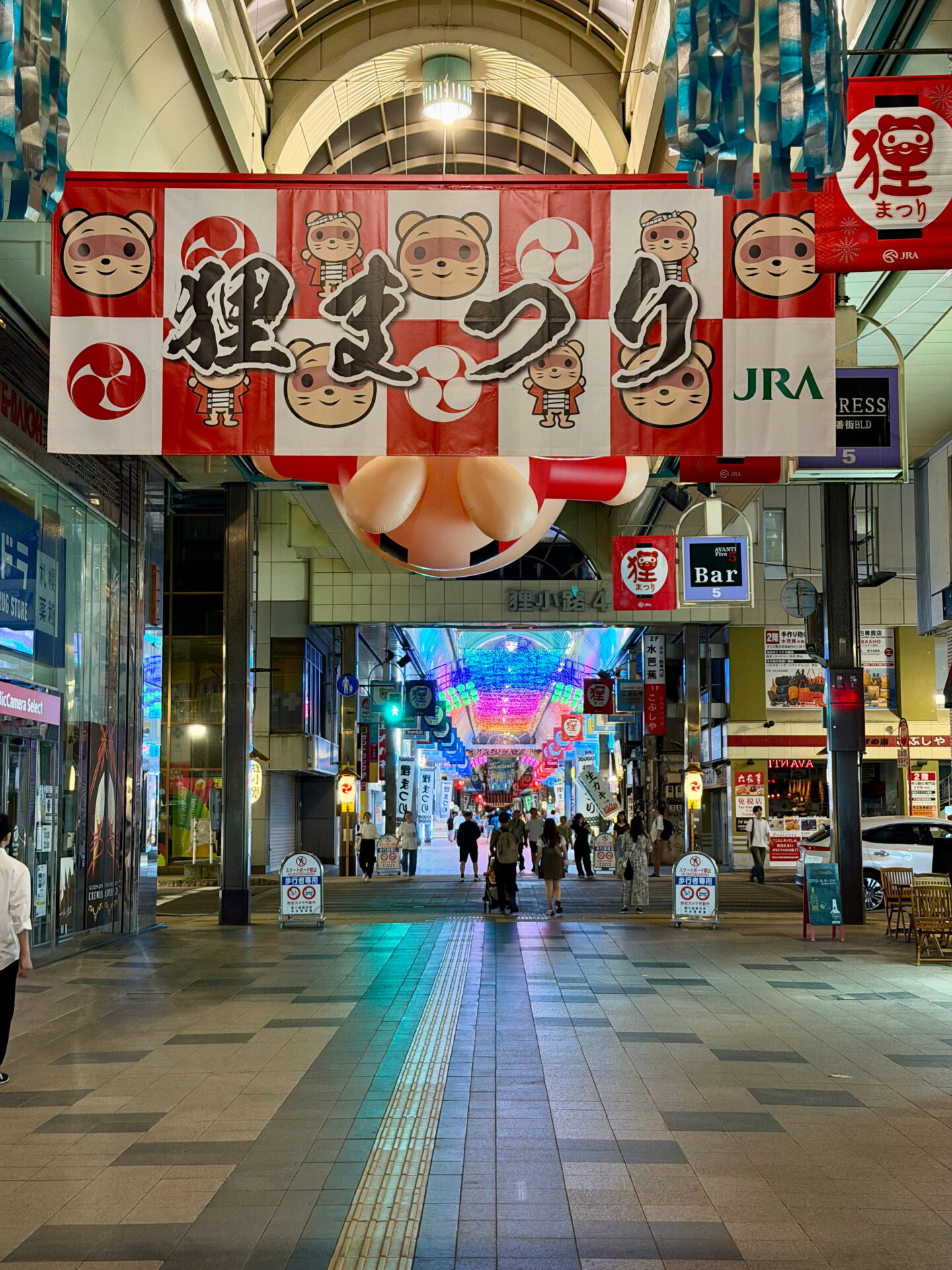 A large red and white festival banner decorated with cartoon tanuki characters and Japanese lettering reading Tanuki Festival hangs above Tanukikoji Shopping Arcade in Sapporo. Colourful illuminated decorations stretch along the covered shopping street as visitors walk beneath them.