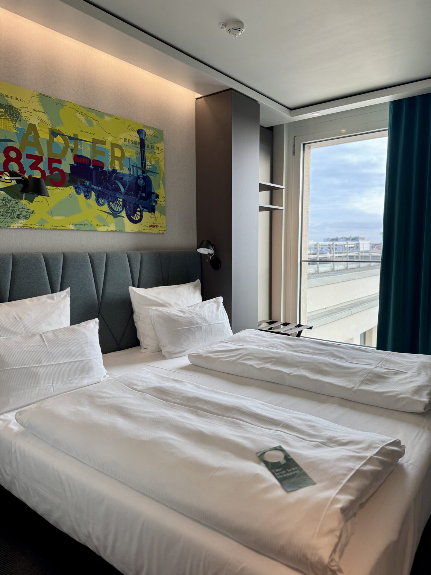 A modern hotel bedroom with two neatly made single duvets on a double bed, white pillows and a grey upholstered headboard. Colourful wall artwork featuring a steam train and the text "ADLER 1835" hangs above the bed, with a large window overlooking nearby buildings.