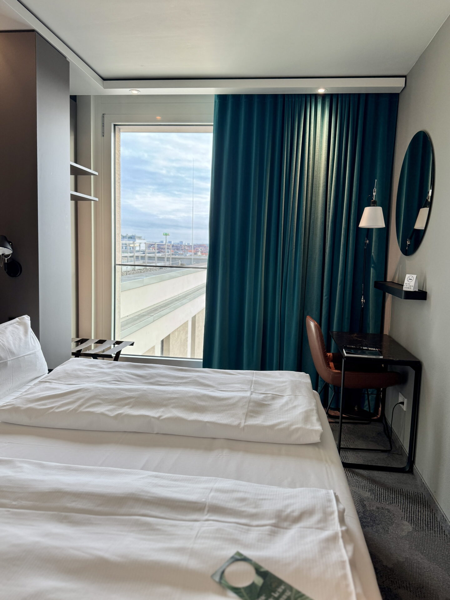 A modern hotel bedroom with white bedding, a large window overlooking the city and floor length teal curtains. A compact black desk, brown chair, wall mounted lamp and round mirror create a small workspace beside the bed.