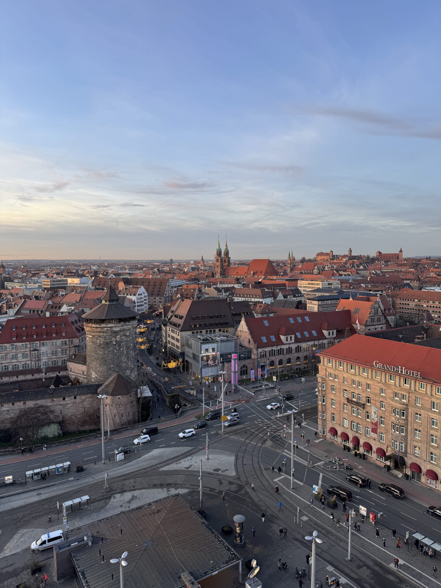 An elevated panoramic view across Nuremberg at sunset, showing historic red tiled rooftops, medieval stone towers and church spires stretching towards the horizon. Roads and tram tracks cross the foreground beside the Grand Hotel, with Nuremberg Castle visible in the distance.