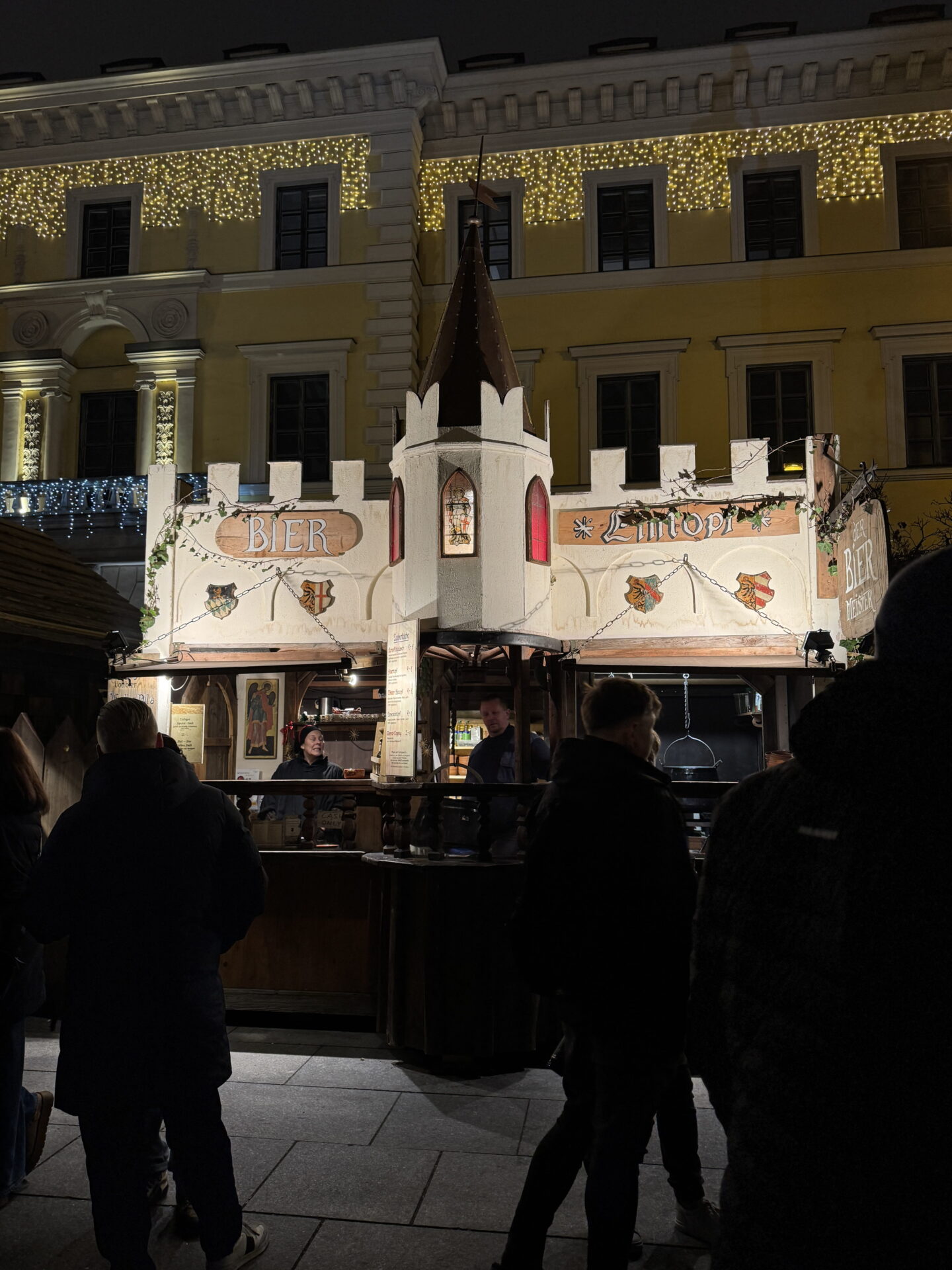 A medieval themed wooden drinks stall at Munich Christmas Market at night, designed like a small castle with white battlements and a pointed central tower. Signs reading "Bier" and "Eintopf" decorate the stall while visitors queue beneath festive lights.