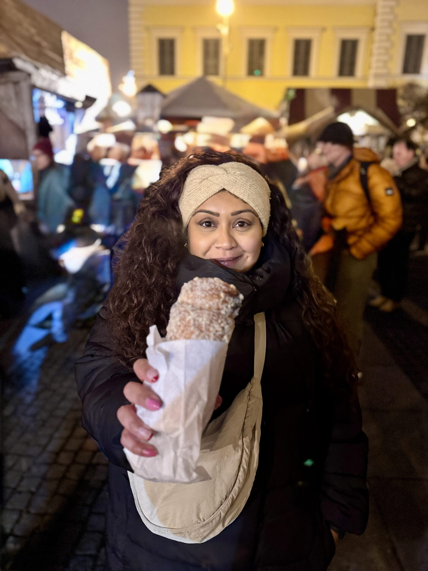 Nishi smiles at the camera while holding out a large sugar coated pastry wrapped in white paper at Munich Christmas Market at night. She wears a cream knitted headband and black puffer coat, with warmly illuminated food stalls and visitors behind her.