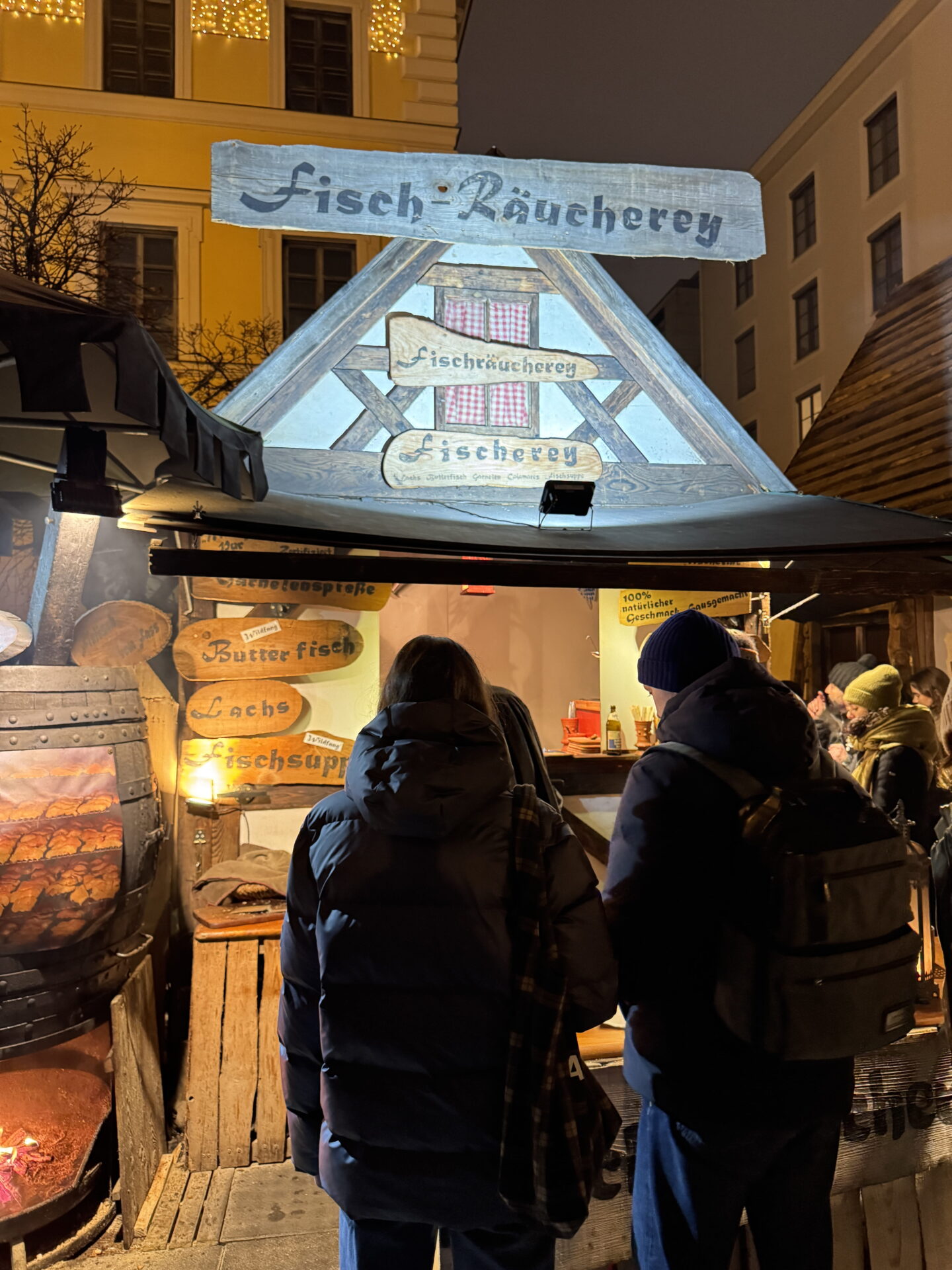 A rustic wooden fish stall at Munich Christmas Market at night, with a glowing smoking oven and customers waiting at the counter. Hand painted signs advertise "Fisch Räucherei", "Fischerei", "Butterfisch", "Lachs" and "Fischsuppe".
