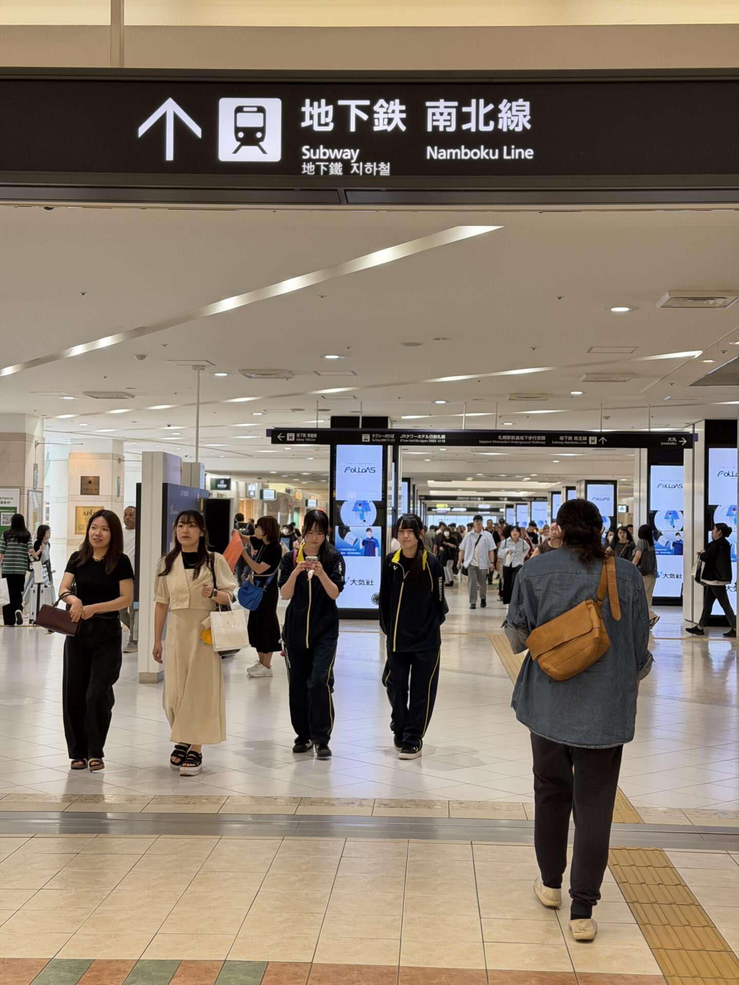 A busy underground shopping concourse in Sapporo filled with pedestrians walking past illuminated advertising screens and shops. An overhead sign reading "Subway Namboku Line" in English and Japanese points straight ahead towards the subway.