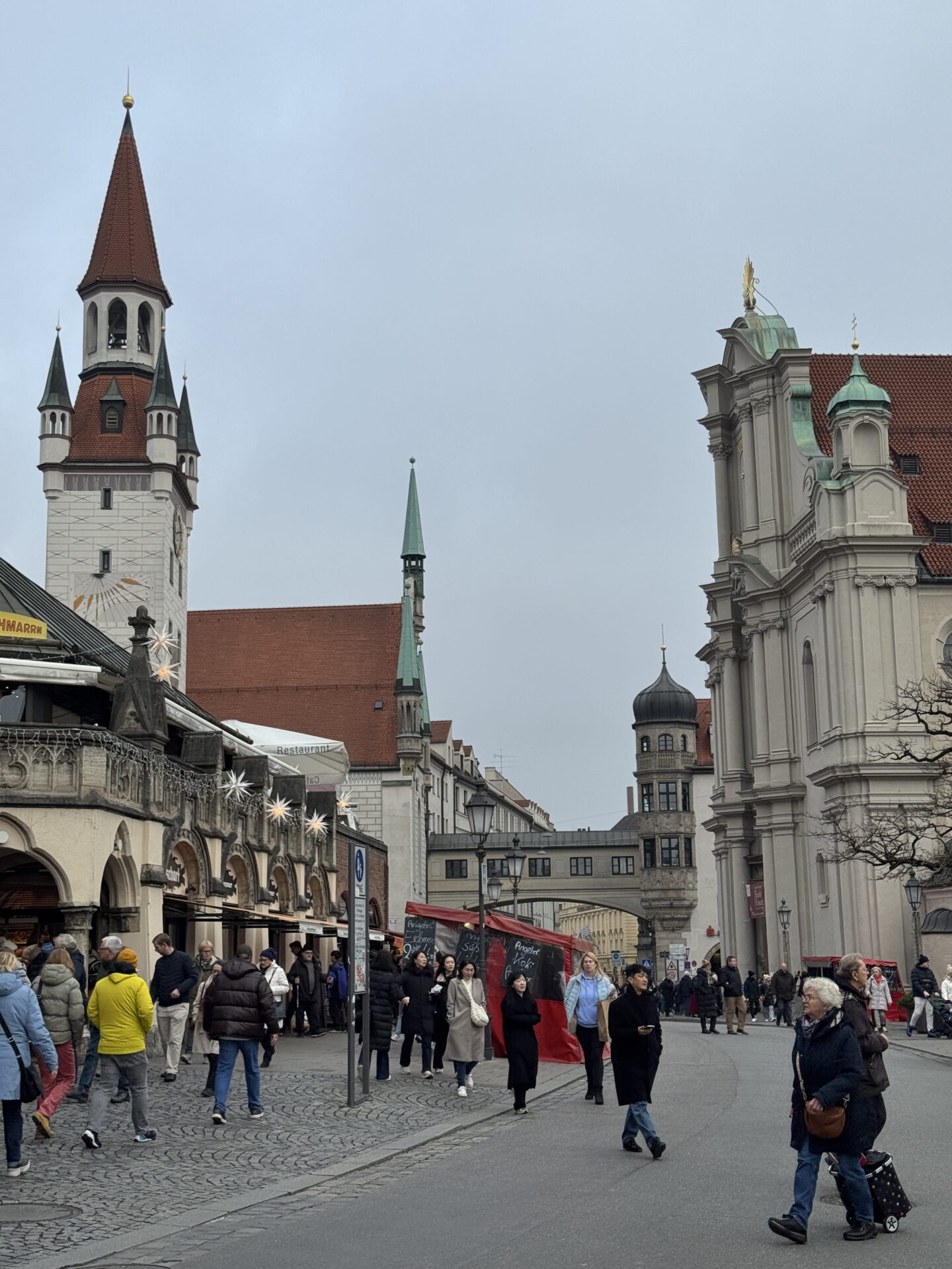 Visitors walking along a cobbled street in Munich Old Town, with historic buildings, pointed church towers and festive star decorations overhead. The white tower of the Old Town Hall and the ornate façade of the Holy Spirit Church frame the street beneath a grey winter sky.