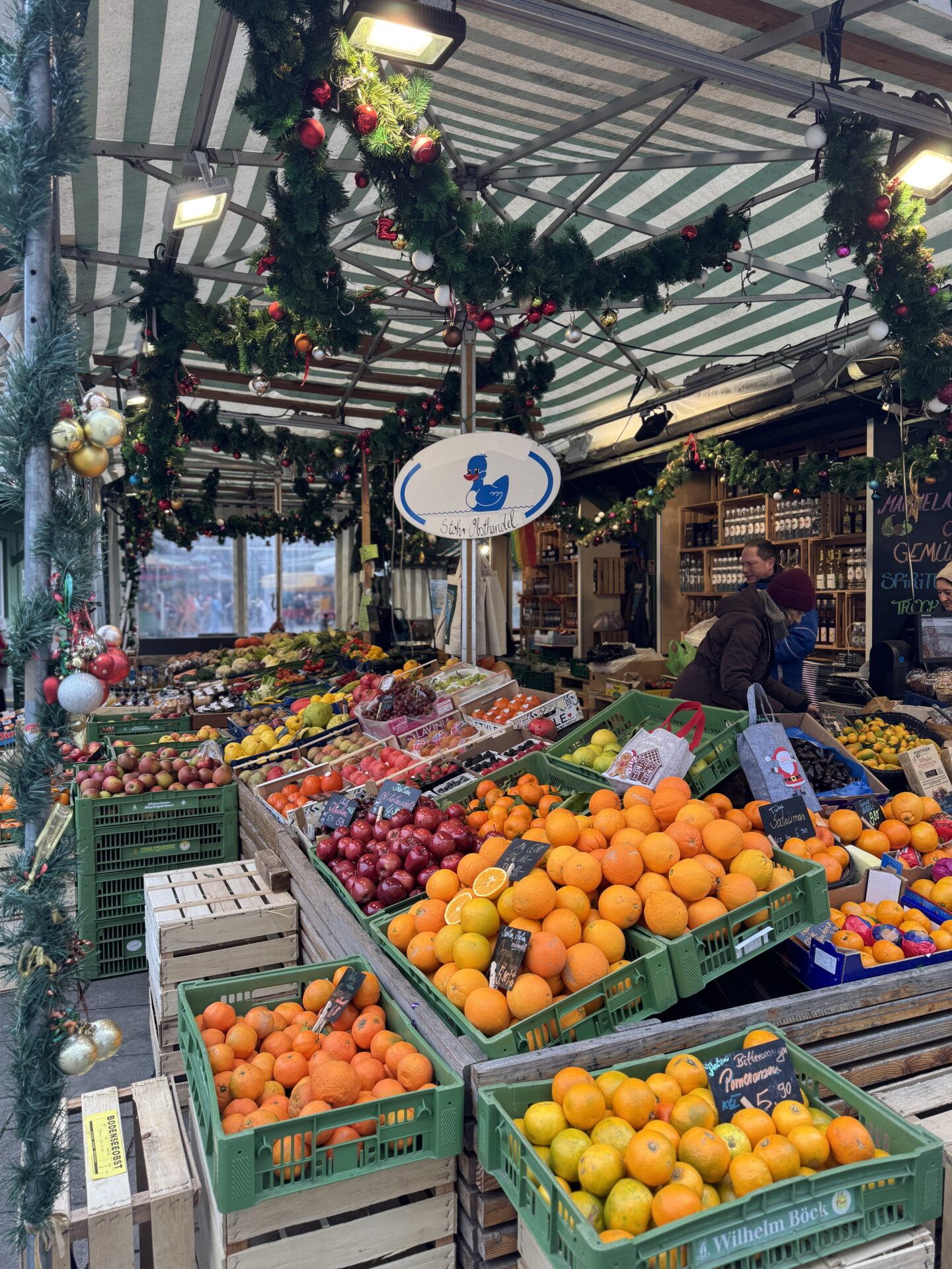 A colourful fruit stall at Munich Viktualienmarkt filled with crates of oranges, mandarins, apples, lemons and other fresh produce. Evergreen garlands decorated with red and gold baubles hang beneath the green and white striped canopy, adding festive touches to the market.