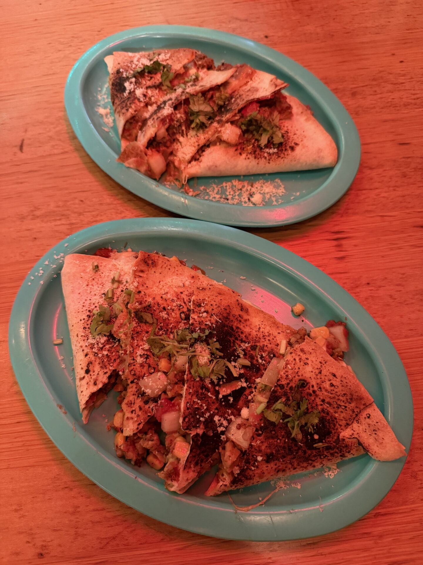 Two turquoise plates of toasted Mexican quesadillas cut into triangular portions and filled with vegetables, sweetcorn and melted cheese. The quesadillas are garnished with chopped coriander, grated cheese and dark seasoning beneath warm reddish restaurant lighting.
