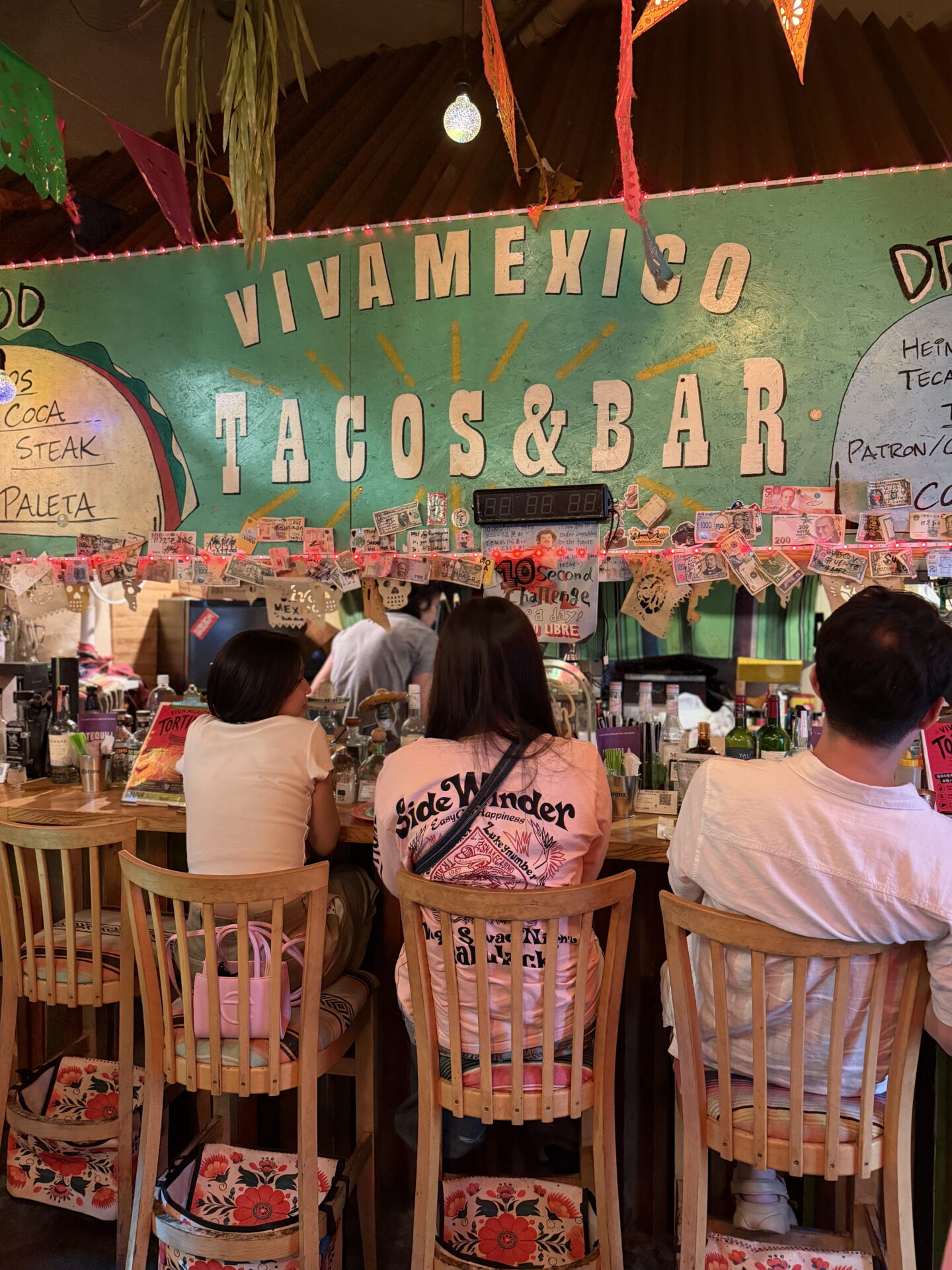 Colourful interior of Viva Mexico Tacos and Bar with a large green wall displaying "VIVA MEXICO TACOS & BAR" above a busy wooden bar counter. Diners sit on tall wooden chairs beneath festive paper decorations, string lights and Mexican themed decorations.