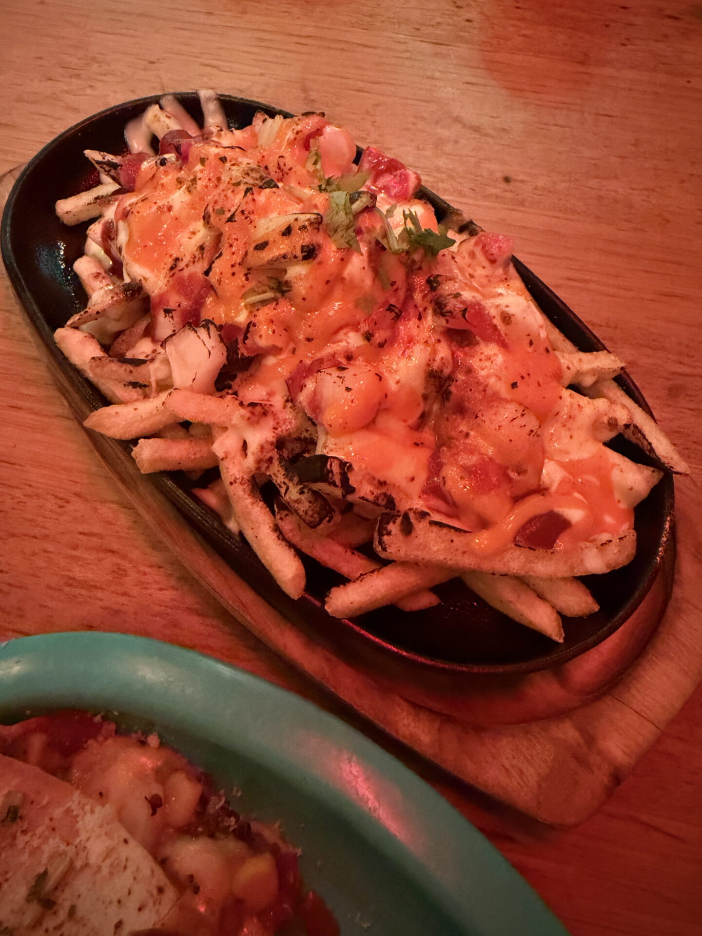 A sizzling black oval platter piled with chips covered in melted orange cheese, diced vegetables, creamy sauce and chopped coriander. The loaded chips are served on a wooden board under warm red restaurant lighting, with part of a turquoise plate visible in the foreground.
