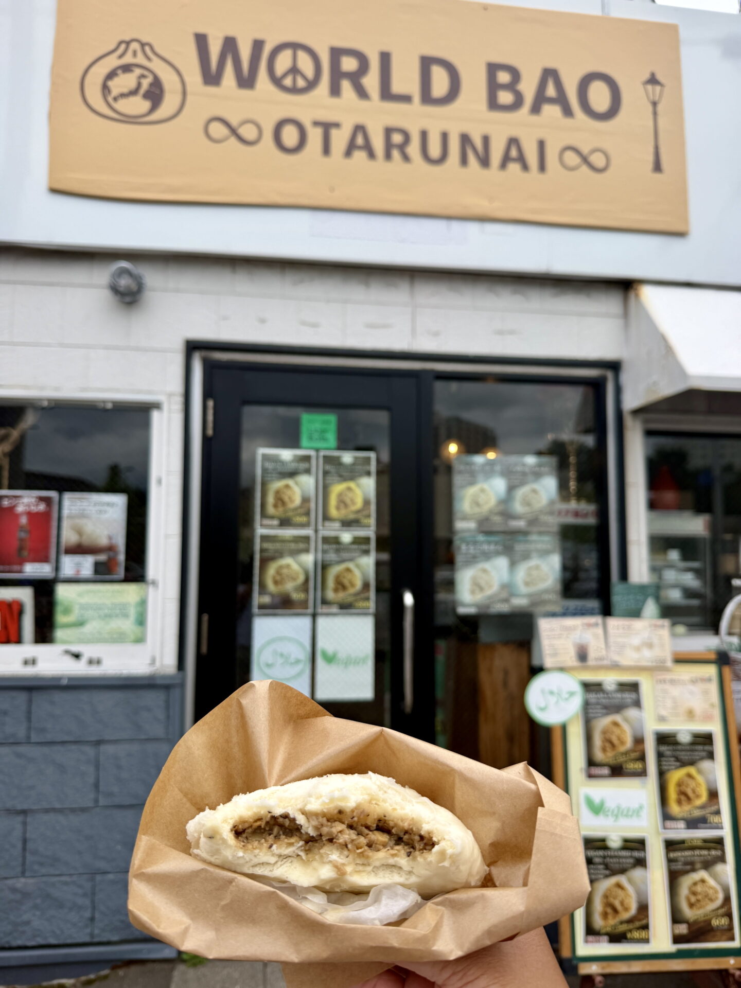 A hand holds a half eaten steamed bao bun filled with minced meat in a brown paper wrapper outside World Bao Otarunai in Otaru. The shop has a large wooden sign reading "WORLD BAO OTARUNAI" above its entrance and photographs of different bao buns displayed on the doors.